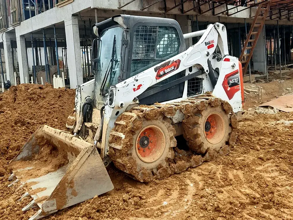 Mini carregadeira Bobcat S570 da Bob Loc com esteira de aço sobre os pneus, em obra no barro